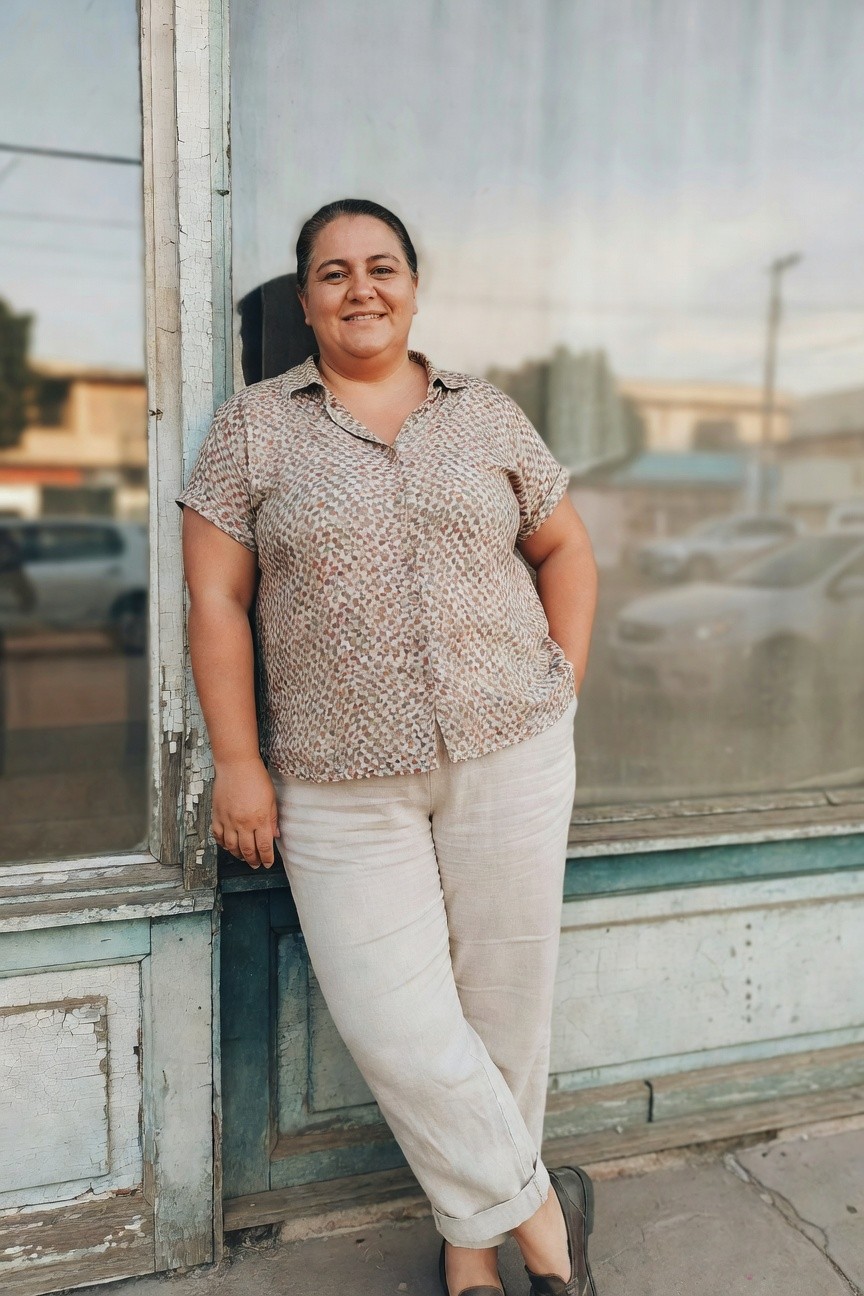 Plus-size woman leaning against a wall in a short-sleeve printed blouse with brown speckles on cream fabric, paired with wide-leg beige linen pants cuffed at the ankles, and brown loafers, hands casually in pockets for a relaxed summer casual look