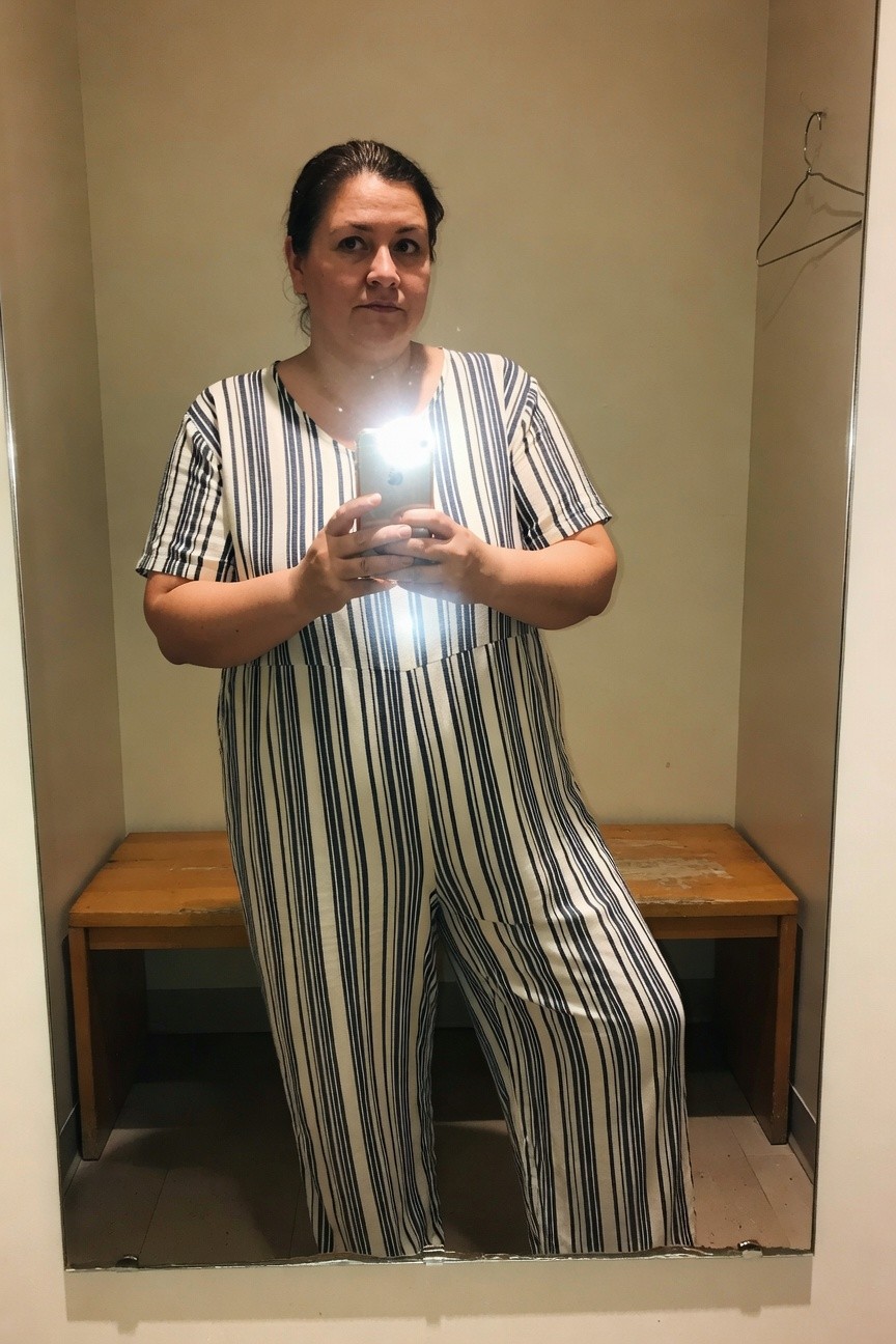Plus-size woman with brown hair taking a mirror selfie in a changing room, wearing a short-sleeve navy and white vertically striped wide-leg jumpsuit with a subtle front pocket detail, standing confidently near a wooden bench