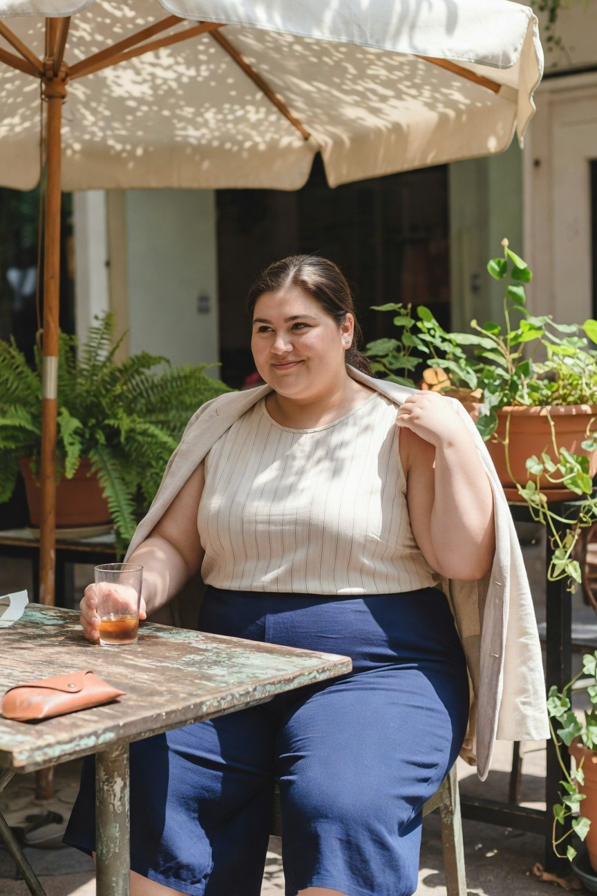 Plus size woman seated outdoors at a wooden table, wearing an open beige linen blazer over a white and beige striped sleeveless tank top, navy wide-leg pants, holding a glass of amber drink, with a brown leather pouch on the table