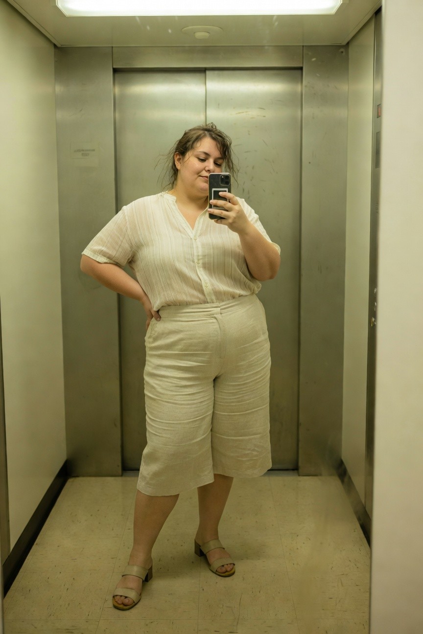 Plus-size woman taking a selfie in an elevator, wearing a loose white striped linen short-sleeve blouse, matching high-waisted wide-leg white linen cropped pants, tan strappy flat sandals, hands on hips and smiling at the camera