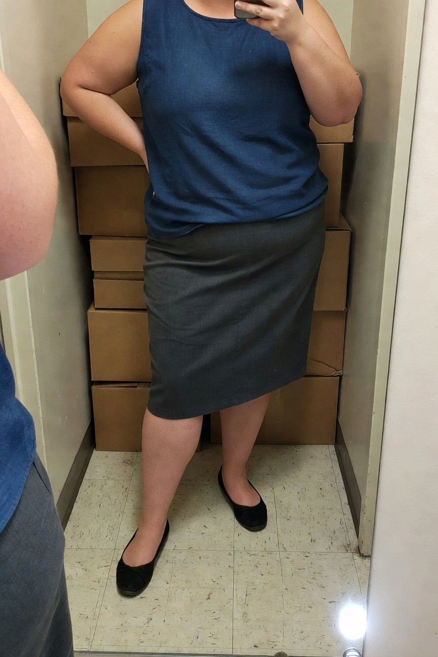 Plus-size woman taking a mirror selfie in a navy sleeveless tank top, gray fitted pencil skirt to the knee, and black bow ballet flats, standing confidently with one hand on hip amid stacked cardboard boxes in a small indoor storage space