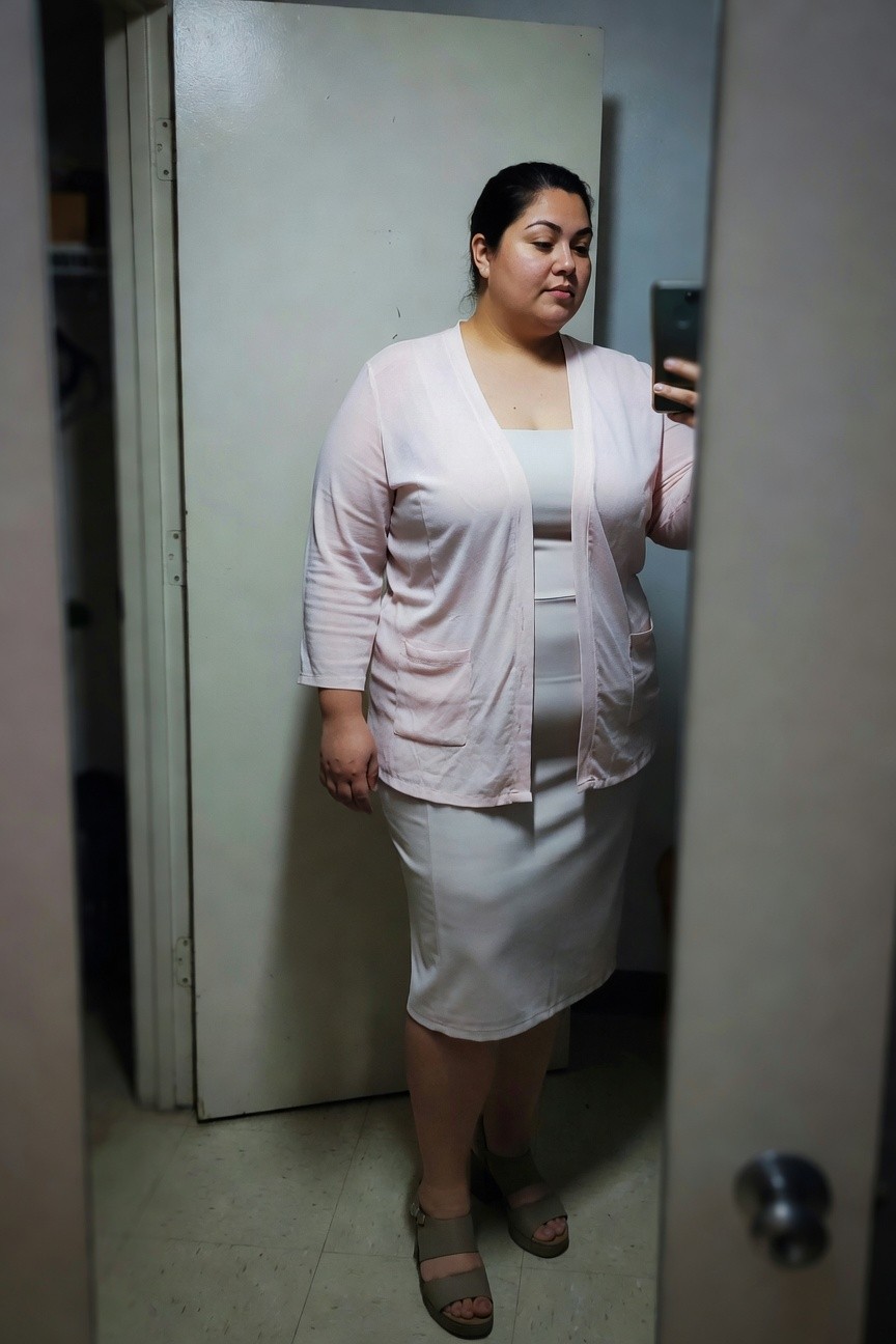 Plus-size woman taking a mirror selfie in a pink open-front cardigan with pockets layered over a white camisole and fitted white sheath dress ending below the knee, wearing tan cork wedge sandals, standing in a closet doorway