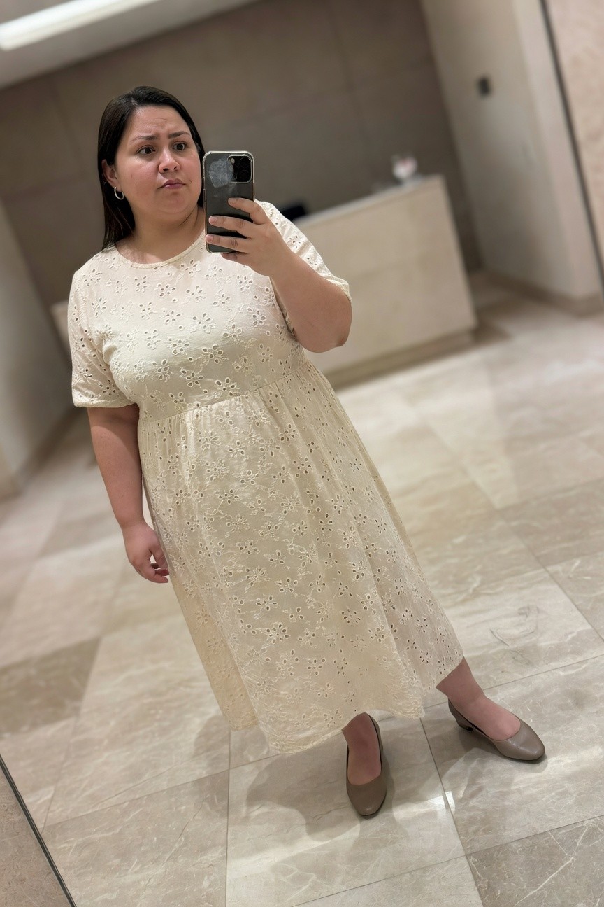 Plus-size Filipina woman with round face and straight dark hair to shoulders, taking a mirror selfie in a white short-sleeved eyelet midi dress with empire waist gather and flared skirt, wearing silver hoop earrings, taupe low-heeled pumps, in a modern marble-floored bathroom lobby.