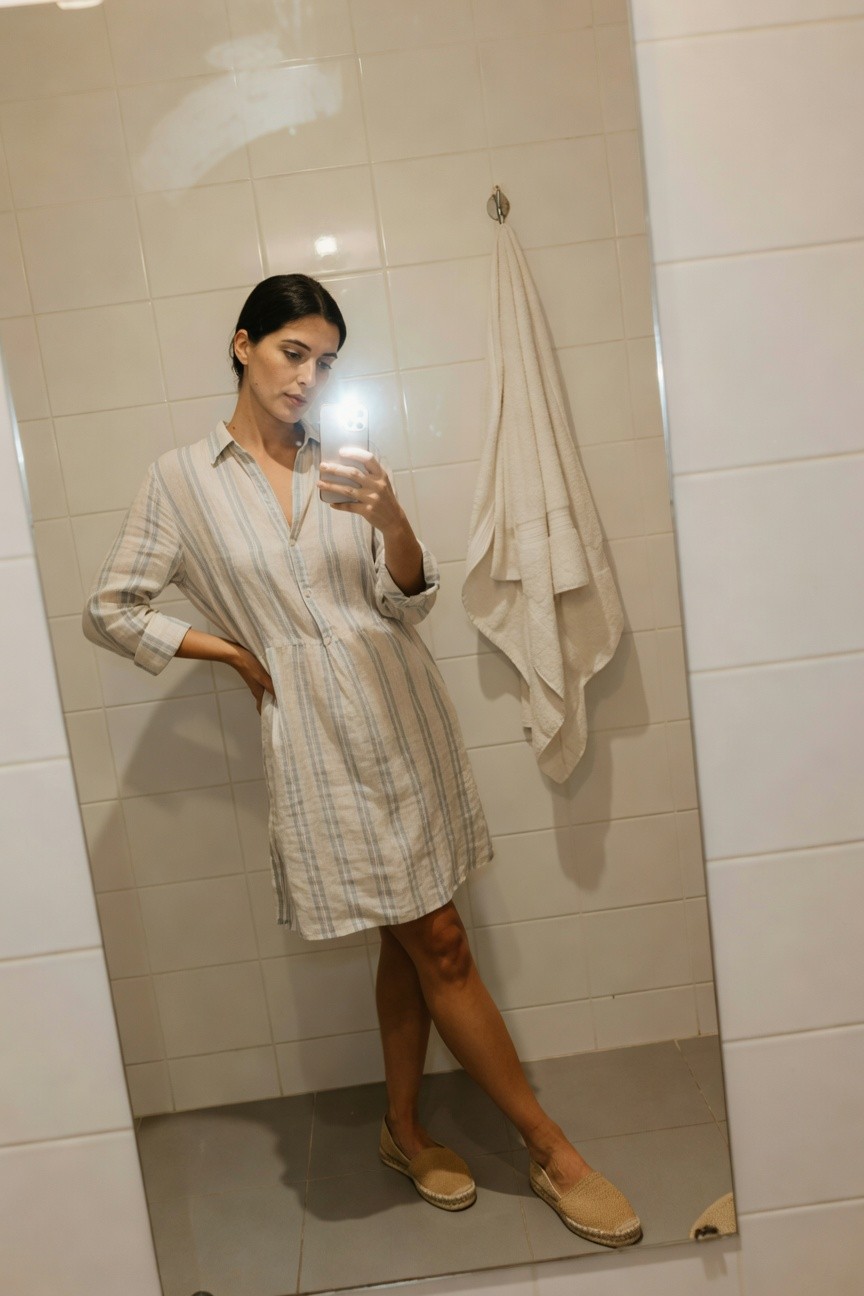 Woman taking a mirror selfie in a bathroom, wearing a light beige striped linen long-sleeve shirtdress with buttons down the front partially open at the collar, sleeves cuffed at elbows, paired with tan espadrilles, one leg slightly bent, white towel hanging nearby
