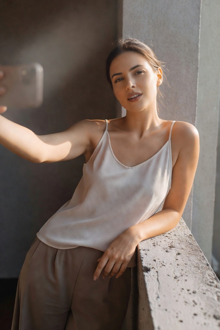 Woman taking a selfie in a white silk spaghetti-strap camisole top and flowing beige wide-leg trousers, leaning casually against a concrete ledge with natural light highlighting the outfit's breezy silhouette