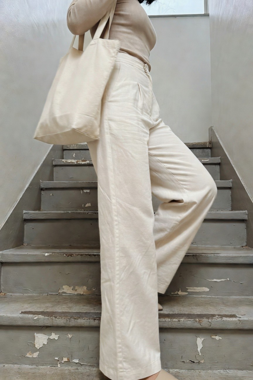 Woman standing sideways on concrete stairs in a fitted cream ribbed knit top, high-waisted cream wide-leg pants, nude slide shoes, and holding a matching cream canvas tote bag