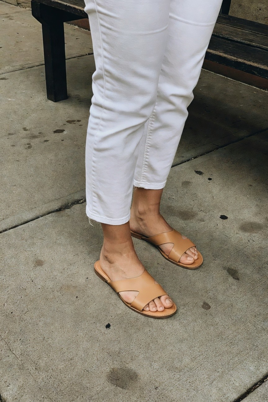 Woman's lower legs in slim white cropped pants cuffed at the ankle and tan leather cross-strap flat sandals, posed near a wooden bench on concrete pavement