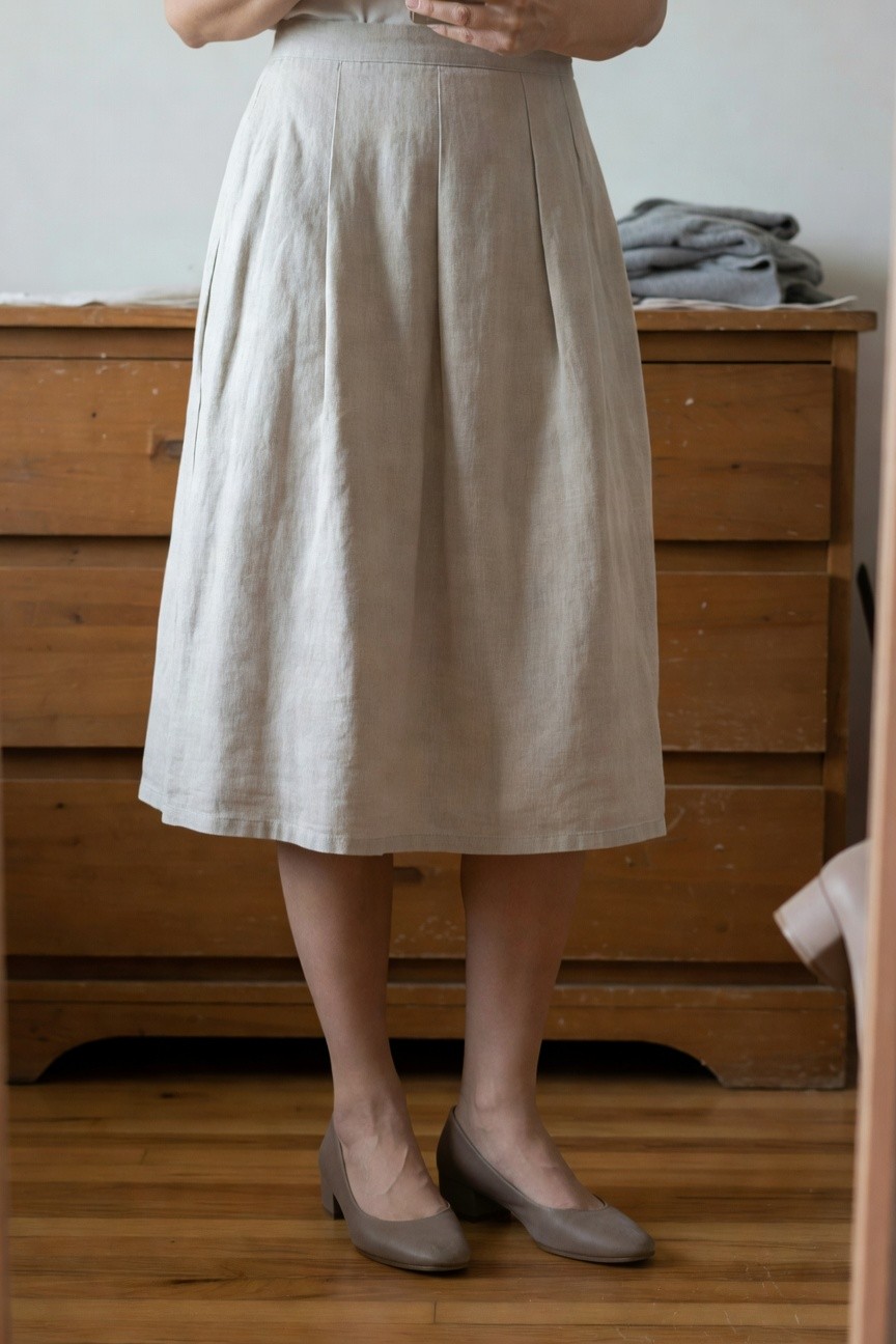 Woman in pale beige pleated linen midi skirt, white sleeveless top, and taupe low block heel pumps, standing near wooden dresser with folded clothes.