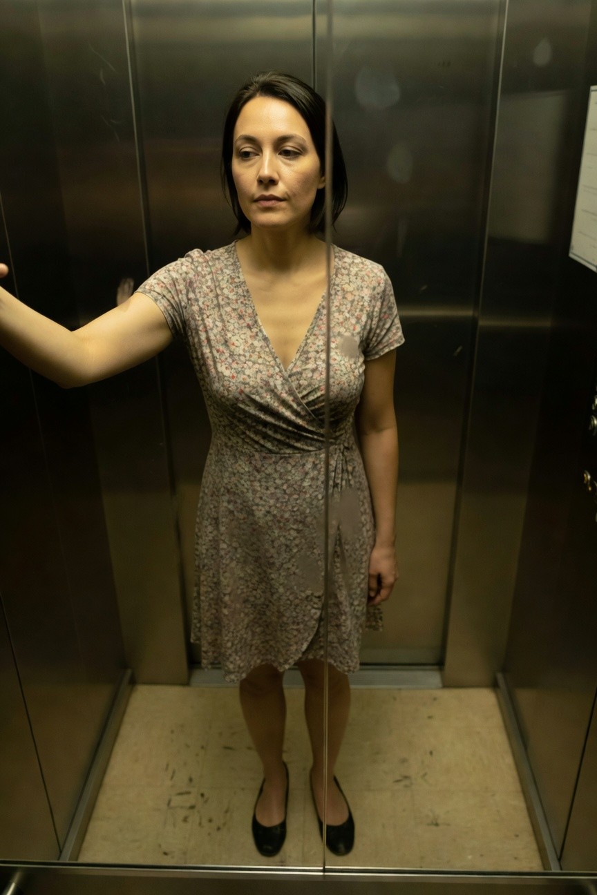 Woman in a short-sleeve floral wrap dress with V-neck and tie belt, black ballet flats, posing with one arm extended in a metallic elevator mirror