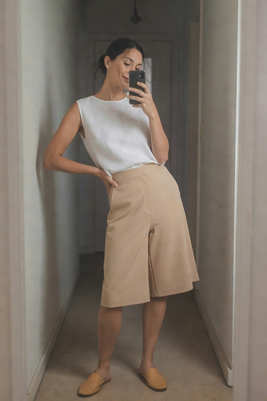 Woman in hallway taking selfie, wearing sleeveless white top, high-waisted beige wide-leg shorts with front slit, and tan slip-on mules, smiling confidently
