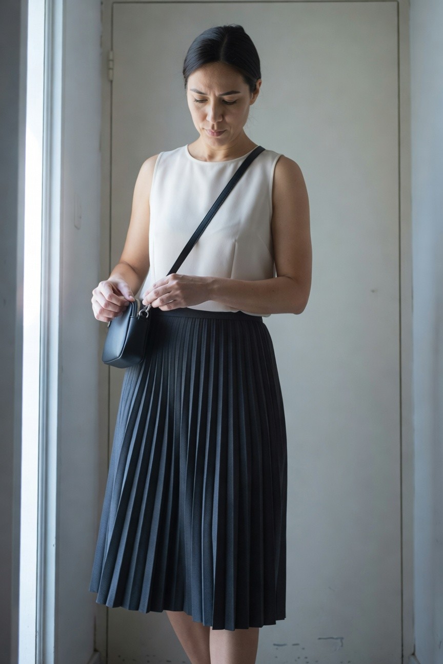 Woman wearing a white sleeveless halter-neck top, black pleated midi skirt, and black crossbody bag, standing by a door with natural light from a window