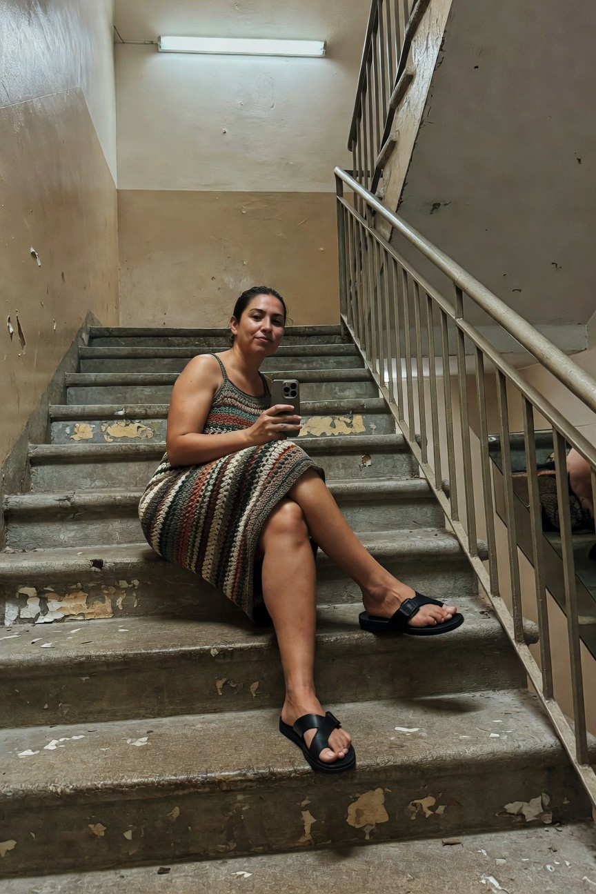 Woman sitting on concrete stairs wearing a sleeveless multicolored striped knit dress in browns greens creams and reds with black strappy flat sandals holding a smartphone selfie style