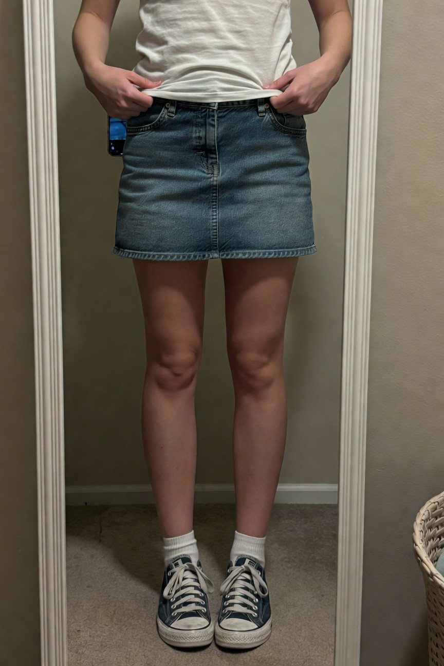 Young woman in full-body mirror selfie wearing a white short-sleeve t-shirt partially tucked into a short light-wash denim mini skirt with hands in pockets and a phone clipped to the waistband, paired with white crew socks and navy low-top Converse sneakers on carpeted floor