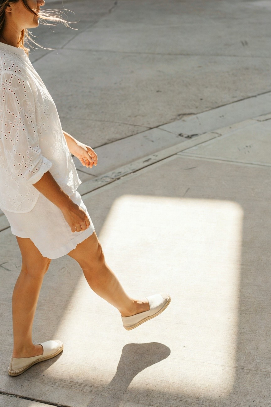 Side view of a woman walking in a white eyelet blouse, matching white linen shorts, and white espadrilles on a concrete surface