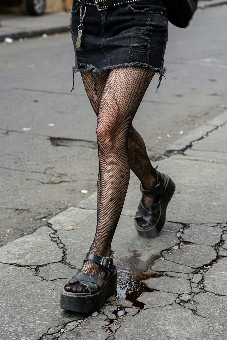 Closeup of a woman's lower outfit featuring a short frayed black denim mini skirt with studded belt and chains, black fishnet stockings, and black platform strappy sandals walking on cracked urban pavement