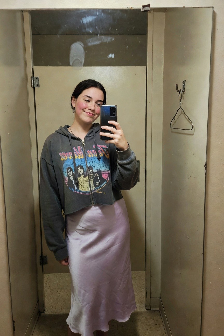 Woman taking mirror selfie in cropped gray zip-up hoodie featuring colorful Doors band graphic layered over pale pink satin slip dress, standing in tiled bathroom with bare feet