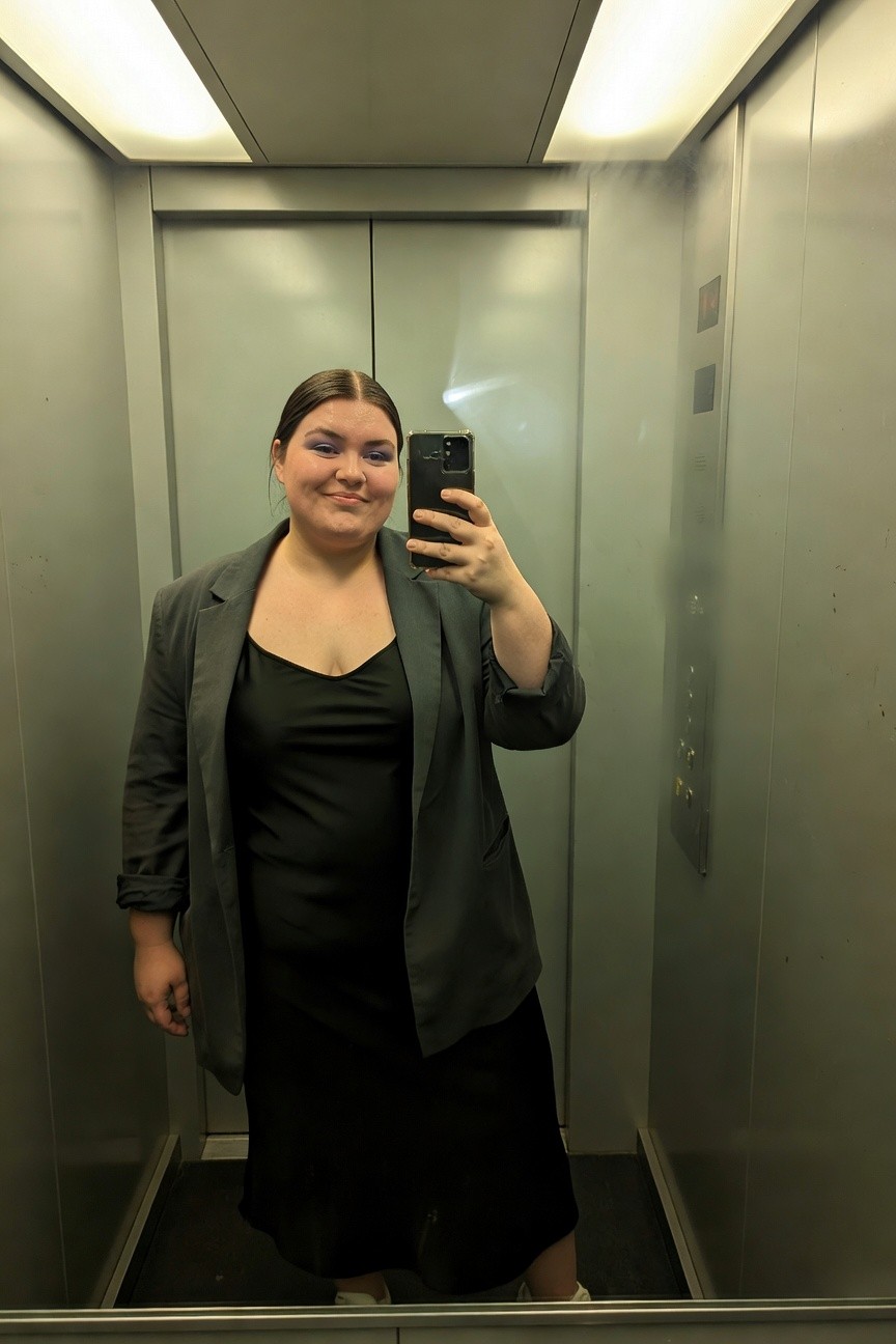 Plus-size woman smiling in selfie wearing black slip maxi dress layered under loose gray blazer with rolled sleeves, white sneakers, taken in elevator mirror
