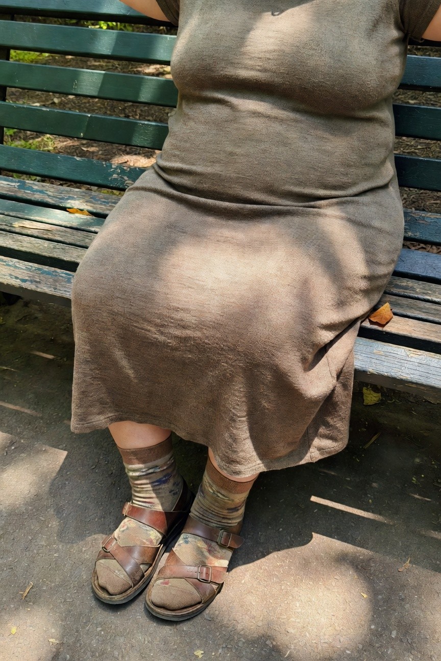 Plus-size woman seated on a wooden park bench in a loose tan maxi dress with short sleeves, camo-patterned socks, and brown leather strap sandals, legs crossed casually