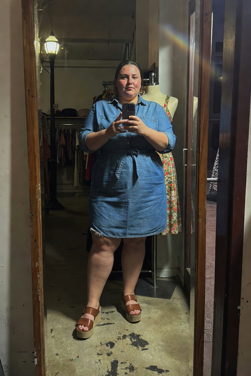 Plus-size woman with round face and flushed cheeks taking a mirror selfie in a clothing store, wearing a blue long-sleeve denim wrap shirt dress with frayed hem and belt over a short colorful floral skirt, tan strappy wedge sandals, standing confidently with hands holding phone.
