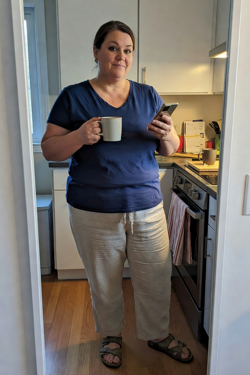 Plus-size woman with brown hair smiling in a kitchen mirror selfie, wearing a navy blue v-neck t-shirt, beige linen drawstring pants, brown leather sandals, holding a white mug in one hand and phone in the other