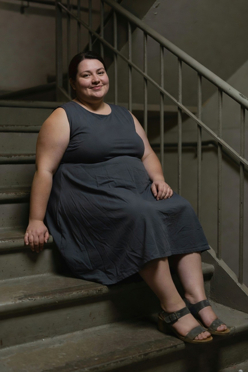 Plus-size woman with dark hair smiling while seated on concrete stairs in a sleeveless charcoal gray A-line tank dress with flared skirt, paired with gray strappy block-heel sandals, metal railing in background
