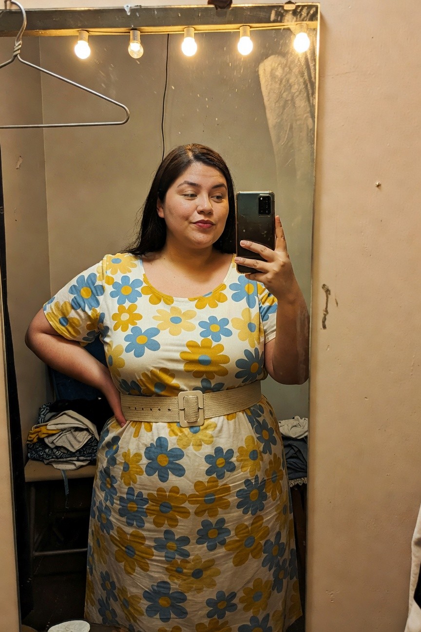 Plus-size woman in a short-sleeve yellow and blue floral print dress with a tan belt at the waist, posing in a dressing room mirror selfie.
