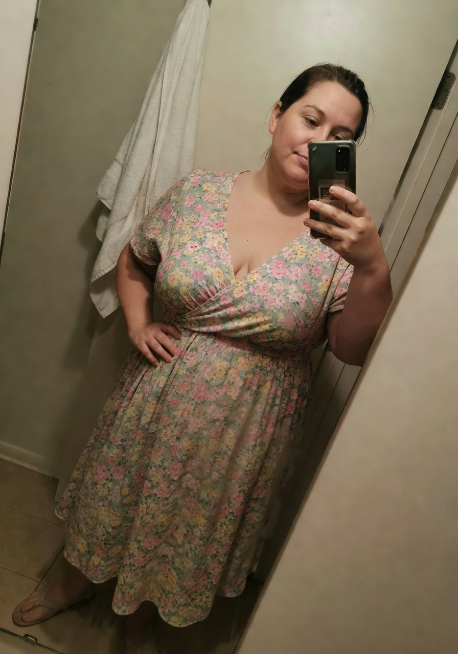 Plus-size woman with dark hair taking a mirror selfie in a light blue floral wrap dress with short sleeves, V-neckline, A-line skirt, hands on hips, wearing tan flip-flop sandals in a bathroom setting