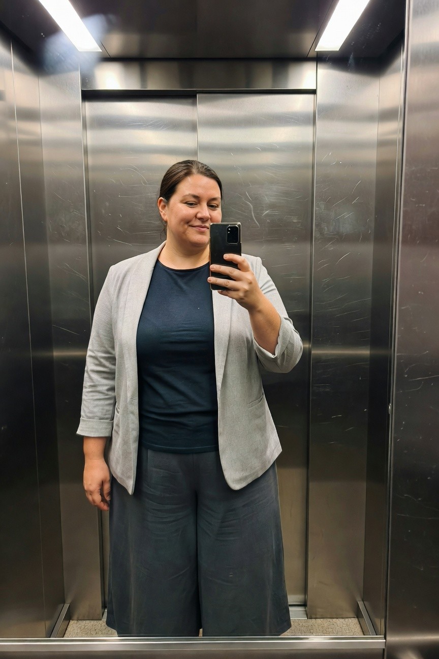 Plus-size woman smiling in elevator selfie wearing open light gray three-quarter sleeve blazer over navy blue crew-neck top and matching gray wide-leg trousers