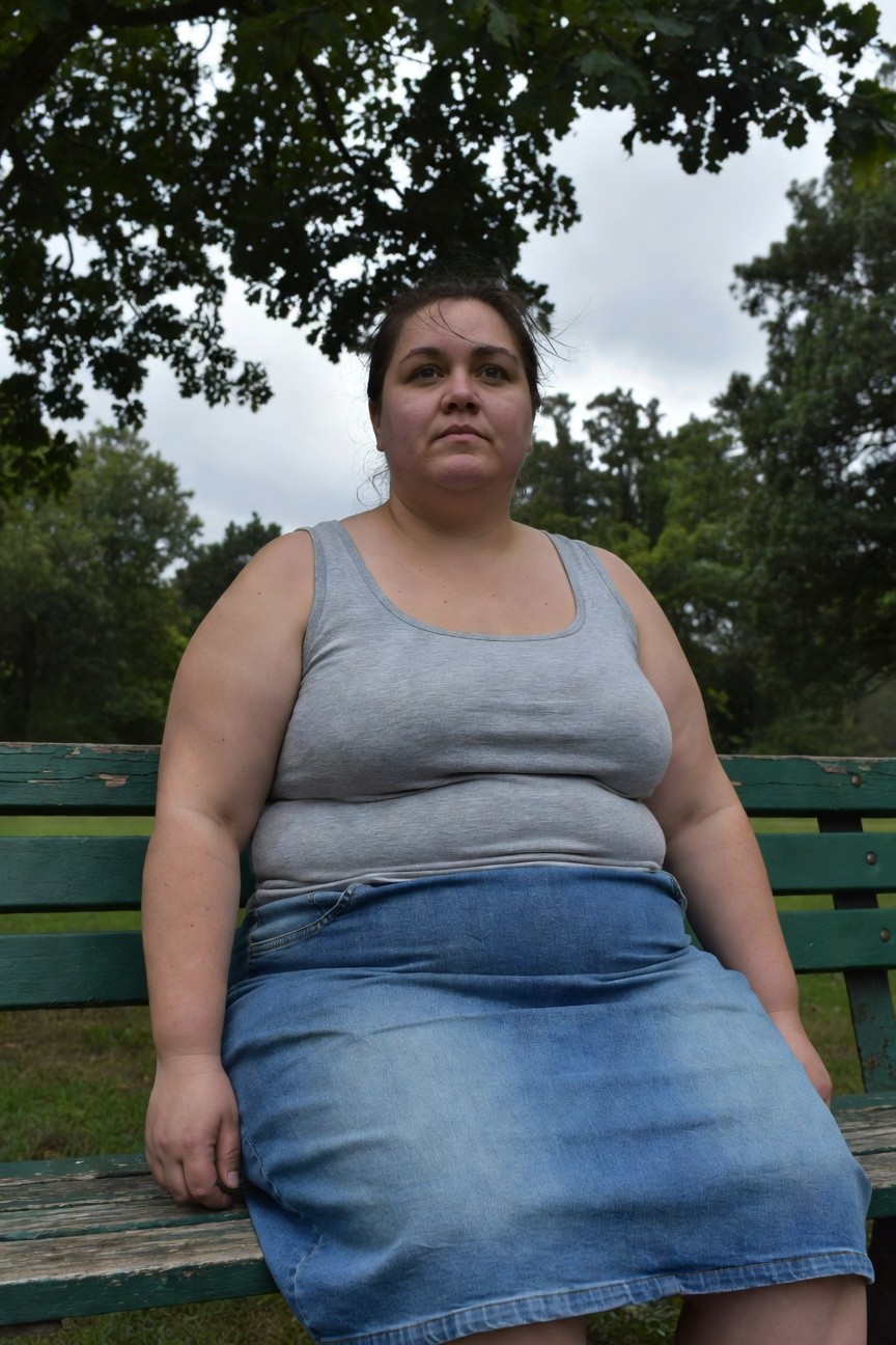 Plus-size woman seated on a green park bench in a gray sleeveless tank top and blue denim skirt, trees in background