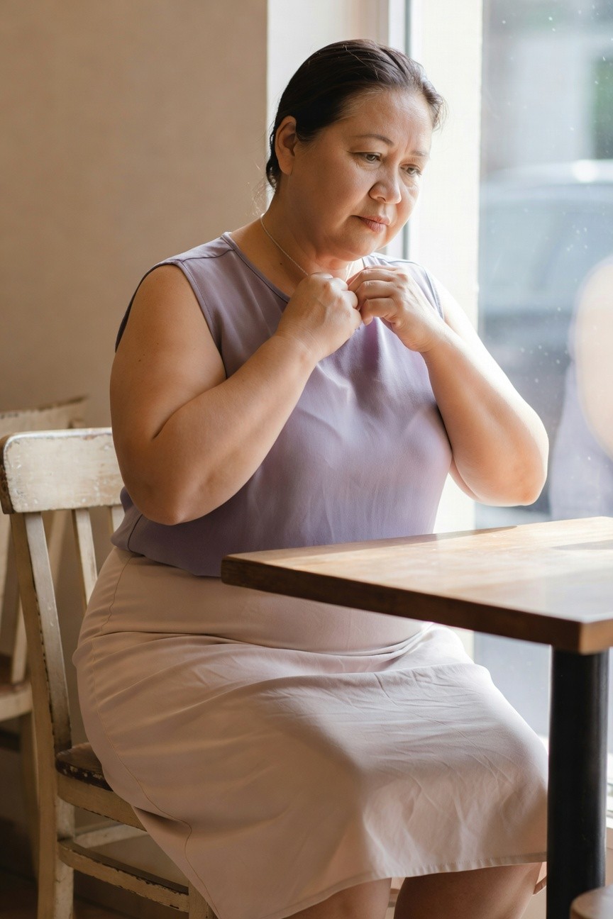 Plus-size Asian woman in her 50s sitting at a wooden cafe table wearing a sleeveless lilac tank top, beige midi skirt, thin necklace, with hands clasped near chest, looking pensively out the window