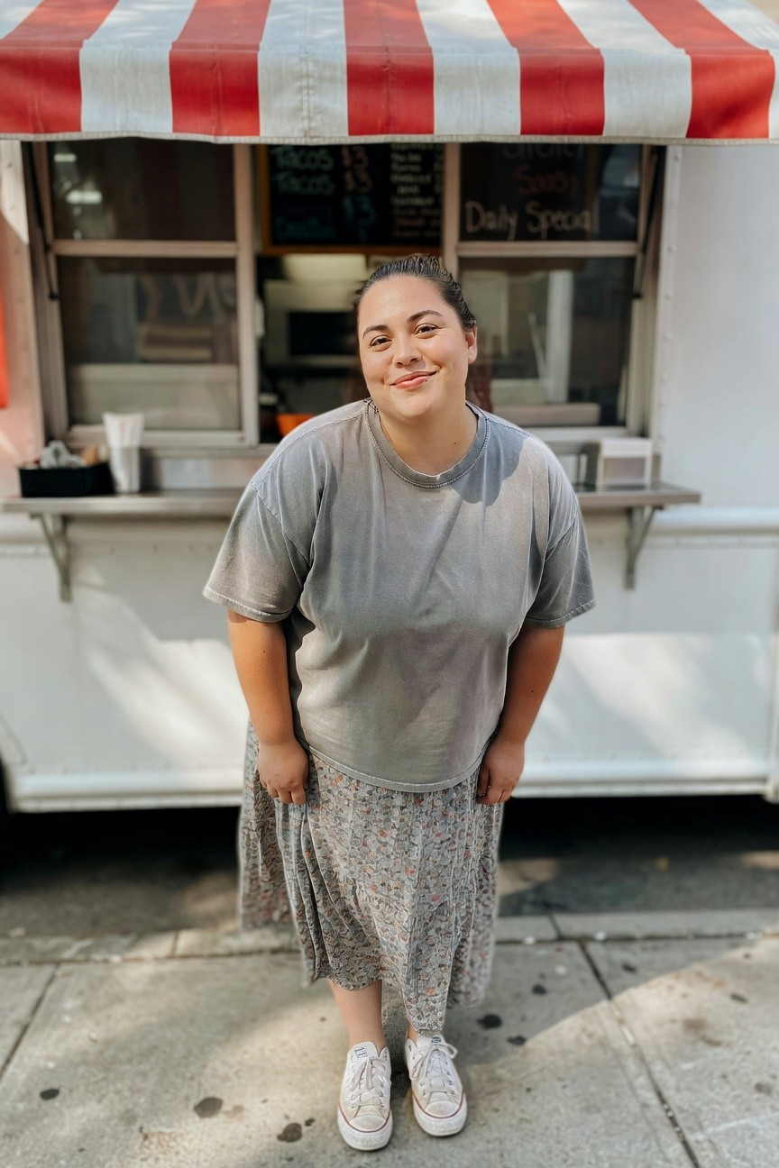 Plus-size woman smiling in front of a food truck, wearing an oversized faded gray t-shirt, tiered floral midi skirt in gray tones with pink and green flowers, and white high-top Converse sneakers, hands in skirt pockets, casual summer outfit.