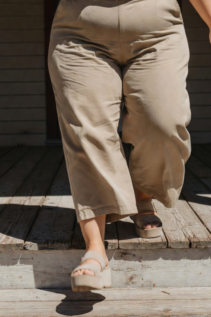 Plus-size woman ascending wooden porch steps in high-waisted beige wide-leg pants and beige platform strappy sandals, emphasizing relaxed summer style