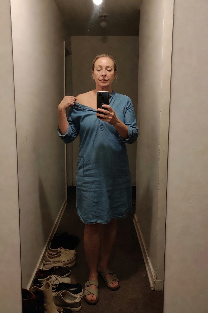 Woman in her 50s with blonde hair taking a mirror selfie in a hallway, wearing a loose blue chambray midi dress with one shoulder slipped down, three-quarter sleeves, and tan gladiator sandals; various shoes scattered on the floor nearby