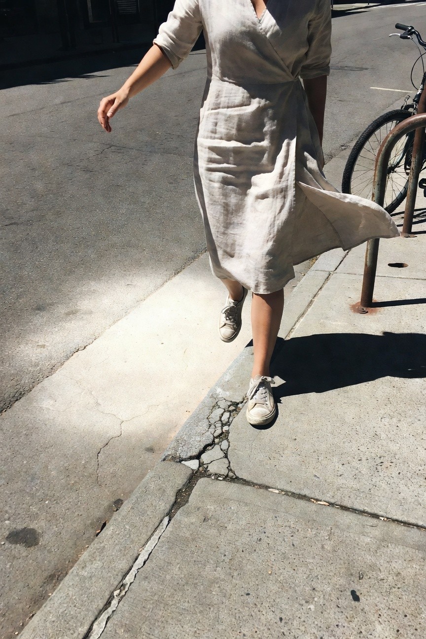 Woman walking in a light beige linen wrap midi dress with three-quarter sleeves and white low-top sneakers on a sidewalk