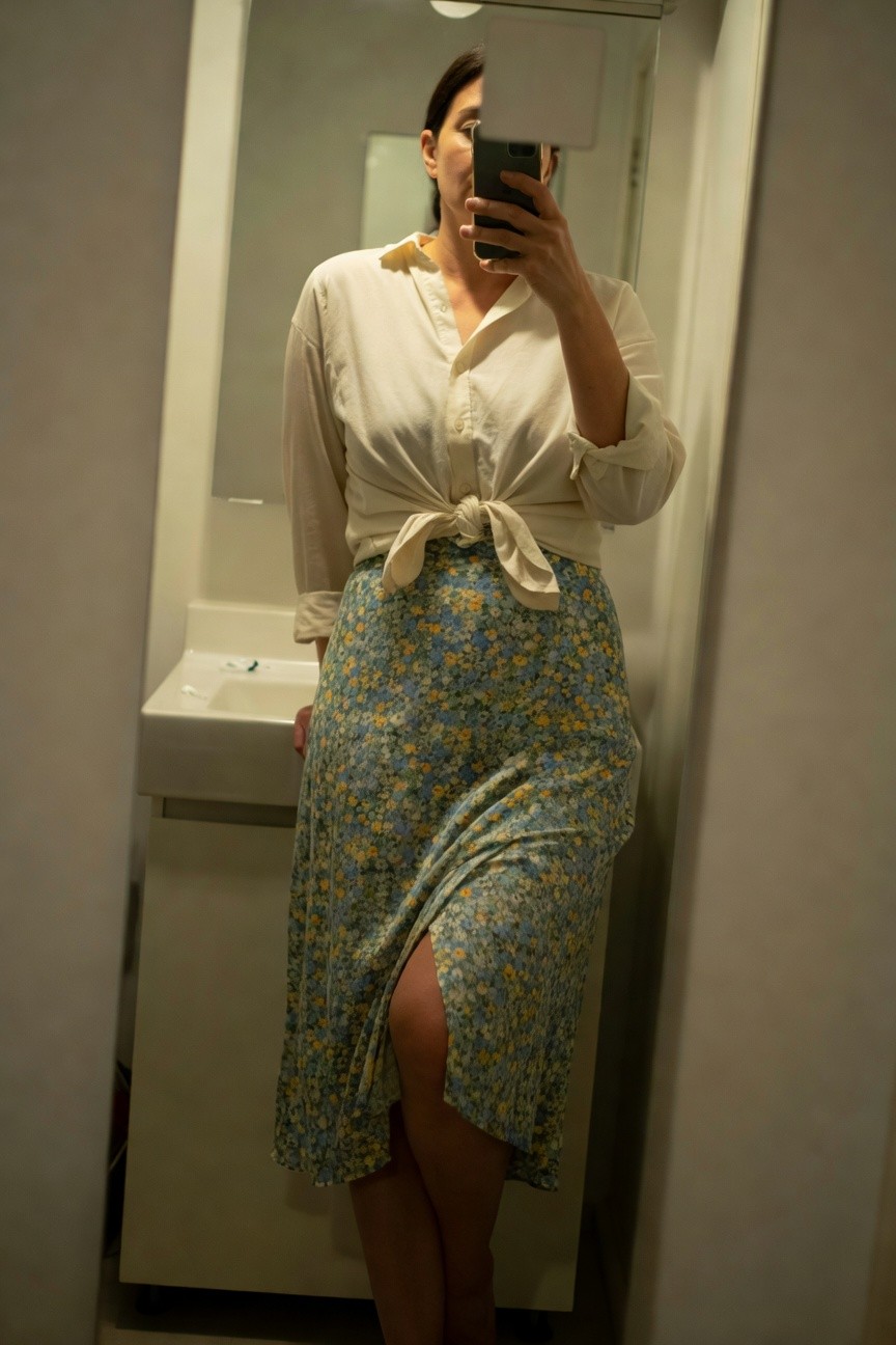 Woman taking mirror selfie in bathroom wearing a white long-sleeve button-up shirt knotted at the waist over a blue-green floral print wrap midi skirt with side slit, standing barefoot by sink.