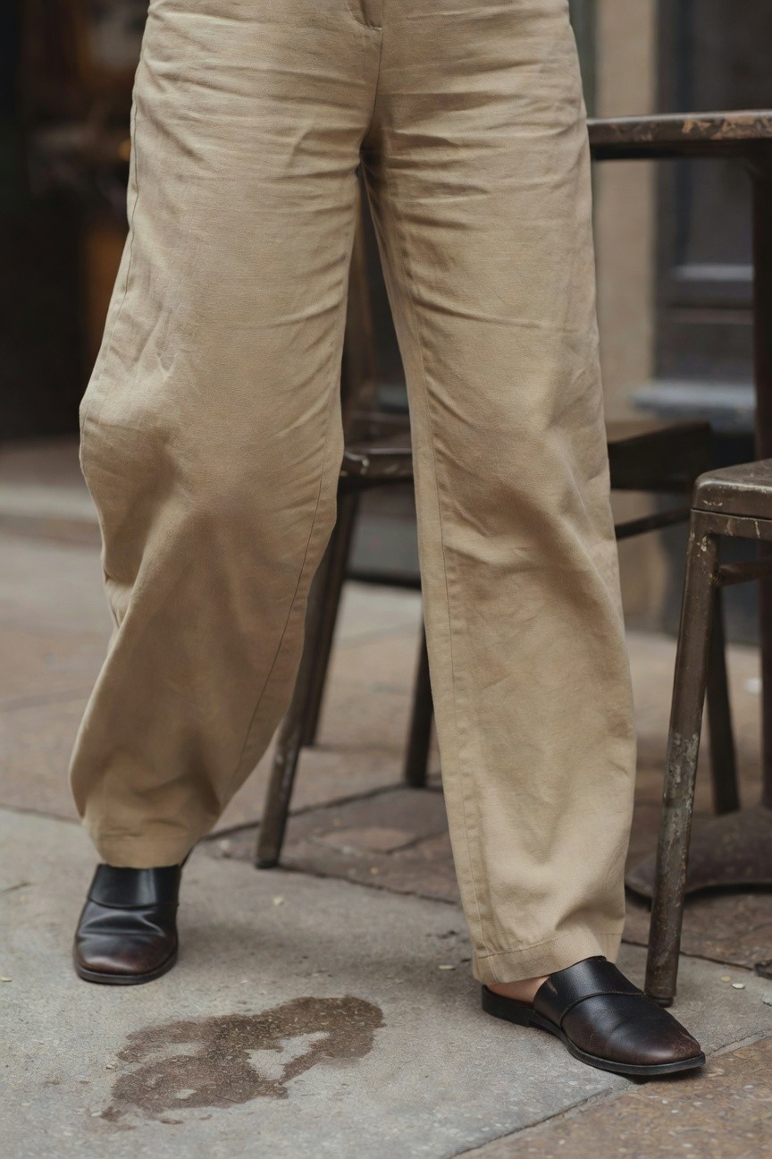 Close-up of beige wide-leg linen pants with natural folds and wrinkles, styled with black leather block-heel mules on a woman standing near metal cafe chairs on a stone pavement