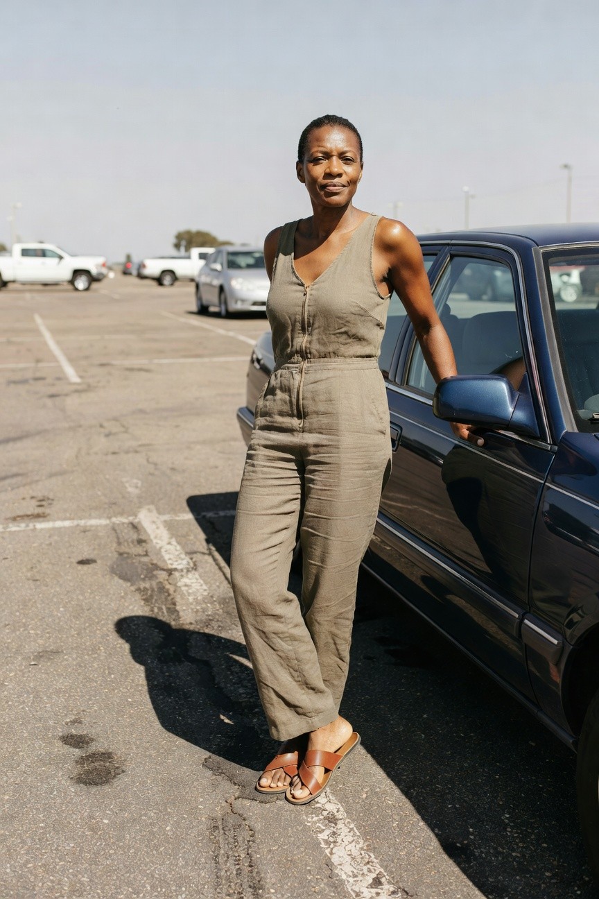 Black woman in sleeveless khaki linen jumpsuit with front zipper wide-leg pants leaning arm on car door wearing tan crossbody bag and brown strappy flat sandals short curly hair neutral expression parking lot setting