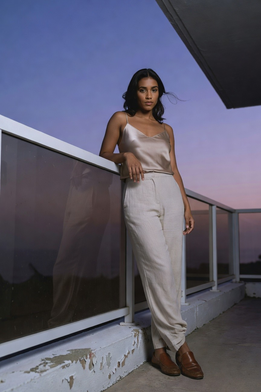 Black woman with wavy dark hair leaning casually on a glass balcony railing, wearing a peachy silk camisole top with thin straps, high-waisted off-white linen wide-leg pants, and brown leather loafers, against a modern balcony setting