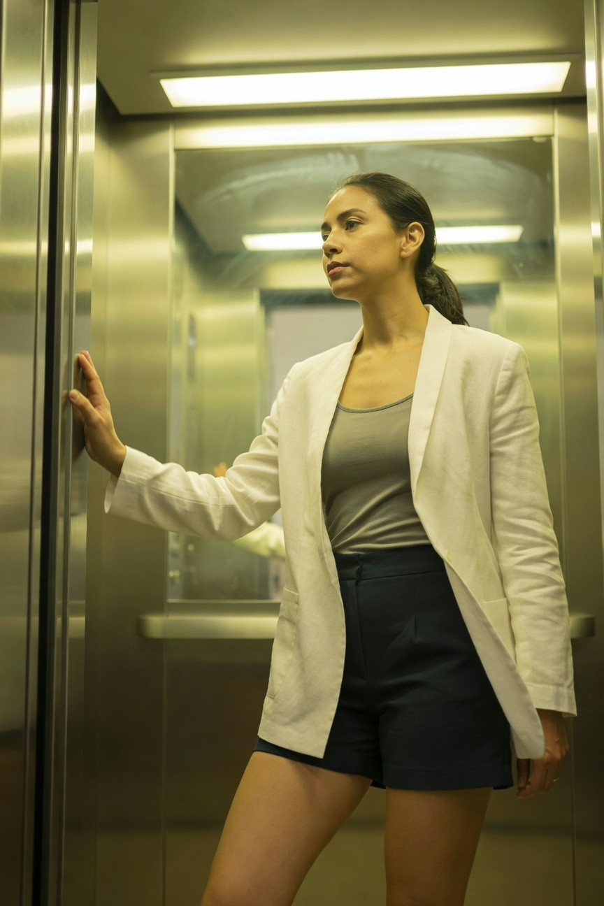 Black woman in open white linen blazer over gray tank top and navy tailored shorts, leaning against elevator wall with one hand, ponytail hairstyle