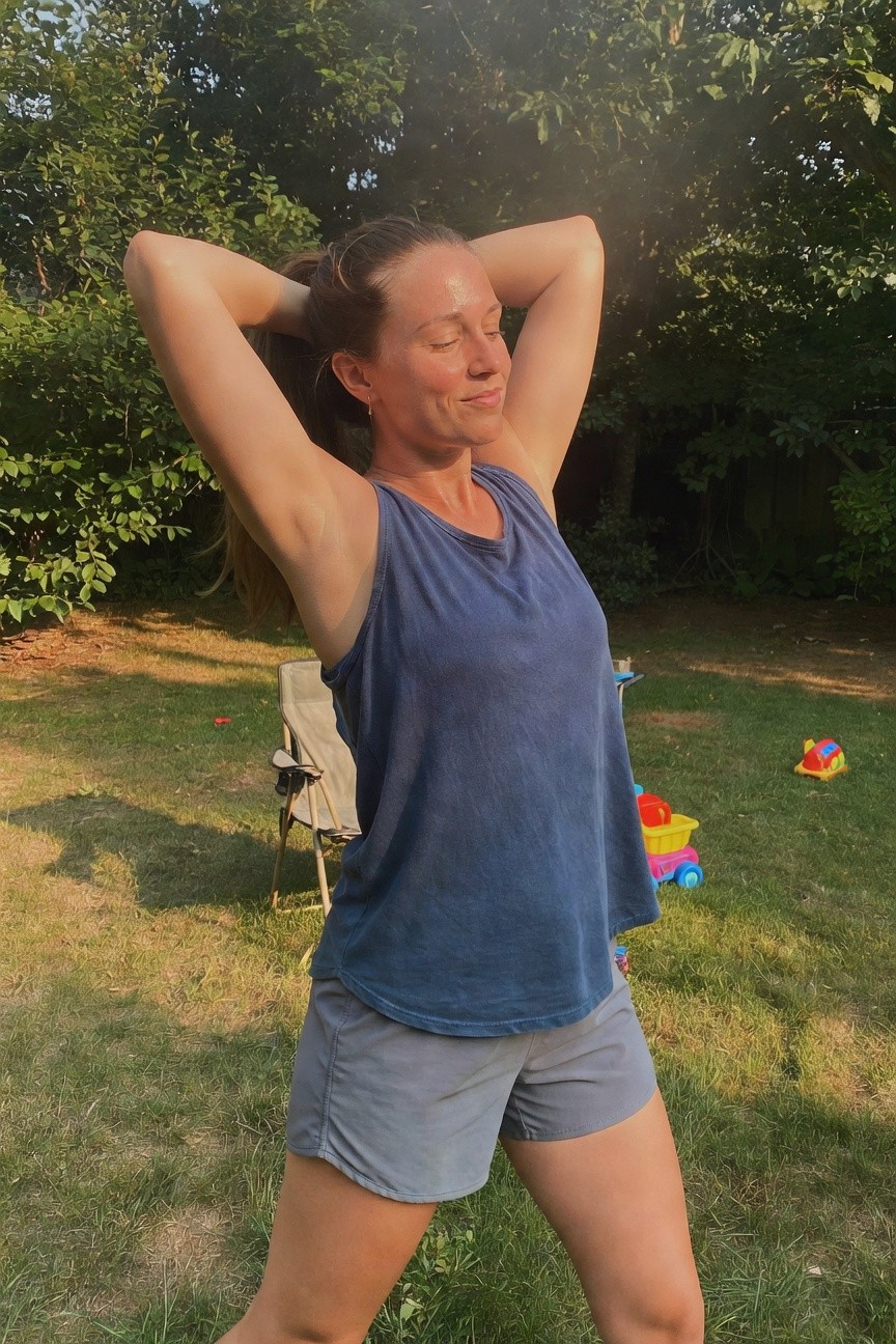 Woman in her 30s stretching arms overhead while smiling, wearing a navy blue sleeveless tank top and gray athletic shorts, standing on grass with backyard chairs and toys in the background