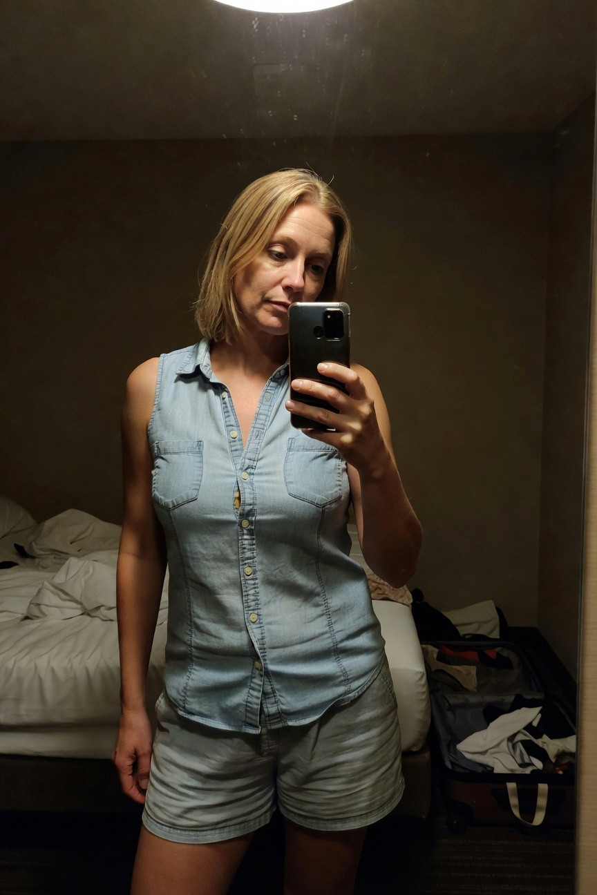 Mirror selfie of a woman in her 40s wearing a light wash sleeveless denim button-up shirt partially unbuttoned over tiny matching denim shorts, standing in a hotel room with bed and suitcase in background