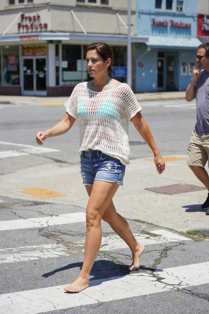 Woman walking barefoot across a crosswalk in a white crocheted open-knit top over a turquoise bikini top and distressed light blue denim cutoff shorts, arm extended while crossing.