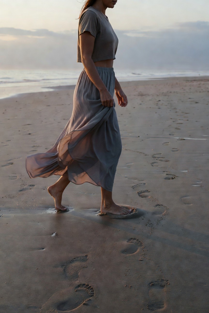 Woman in fitted gray short-sleeve crop top and high-waisted flowy gray midi skirt walking barefoot on sandy beach leaving footprints behind
