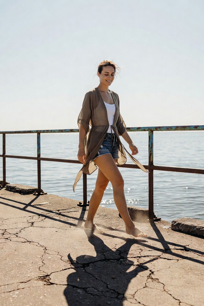 Woman in sheer beige kimono-style jacket over white tank top and cutoff denim shorts, walking barefoot with flowing movement