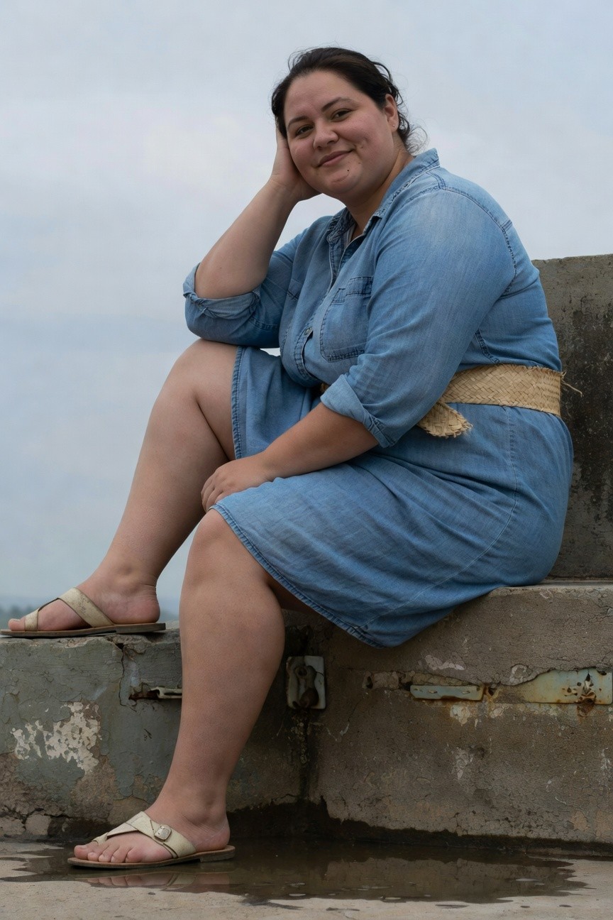 Plus-size woman smiling with hand on cheek, wearing a blue denim shirt dress with rolled sleeves, woven straw belt, and tan flip-flop sandals, sitting on a concrete edge