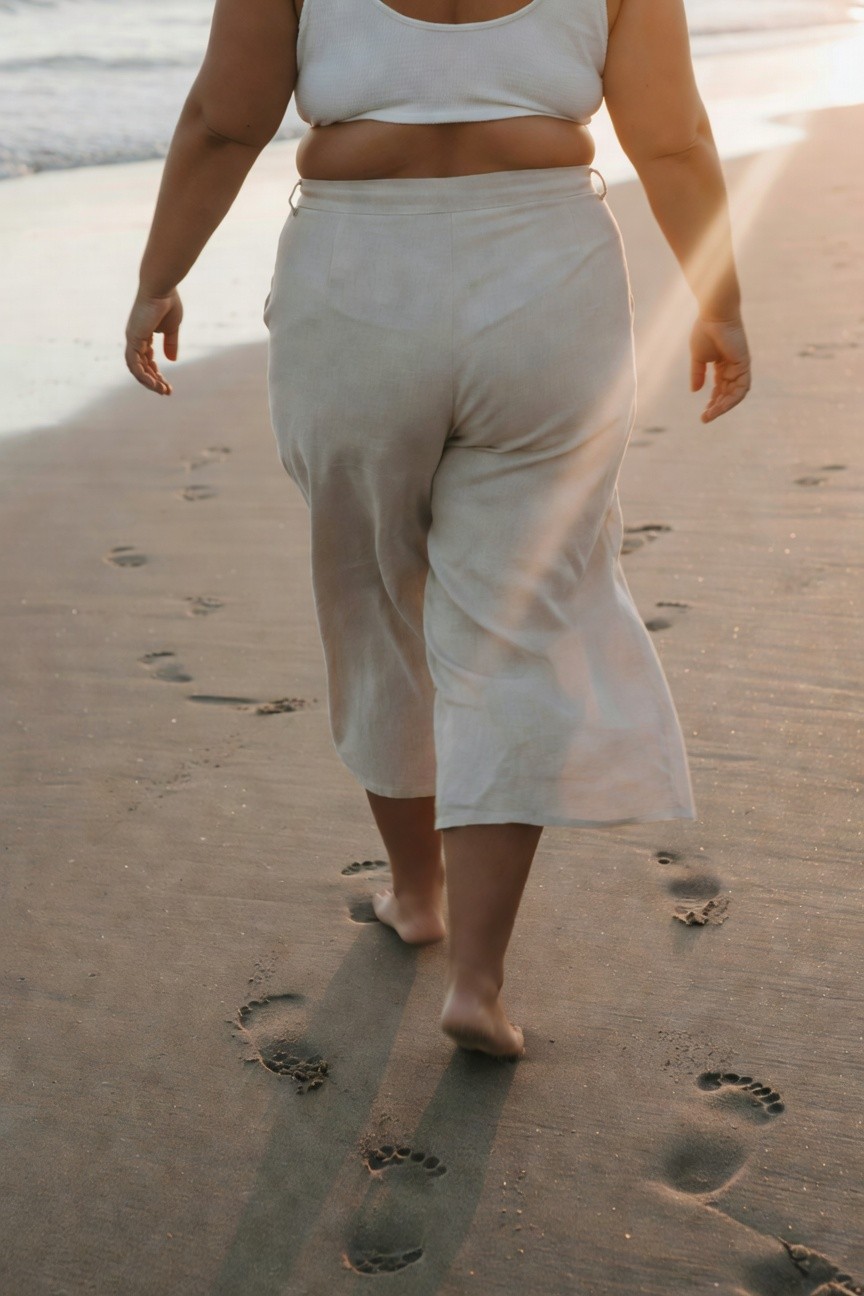 Back view of plus-size woman in white cropped ribbed tank top and high-waisted wide-leg pale linen pants, walking barefoot leaving footprints in sand at sunset beach