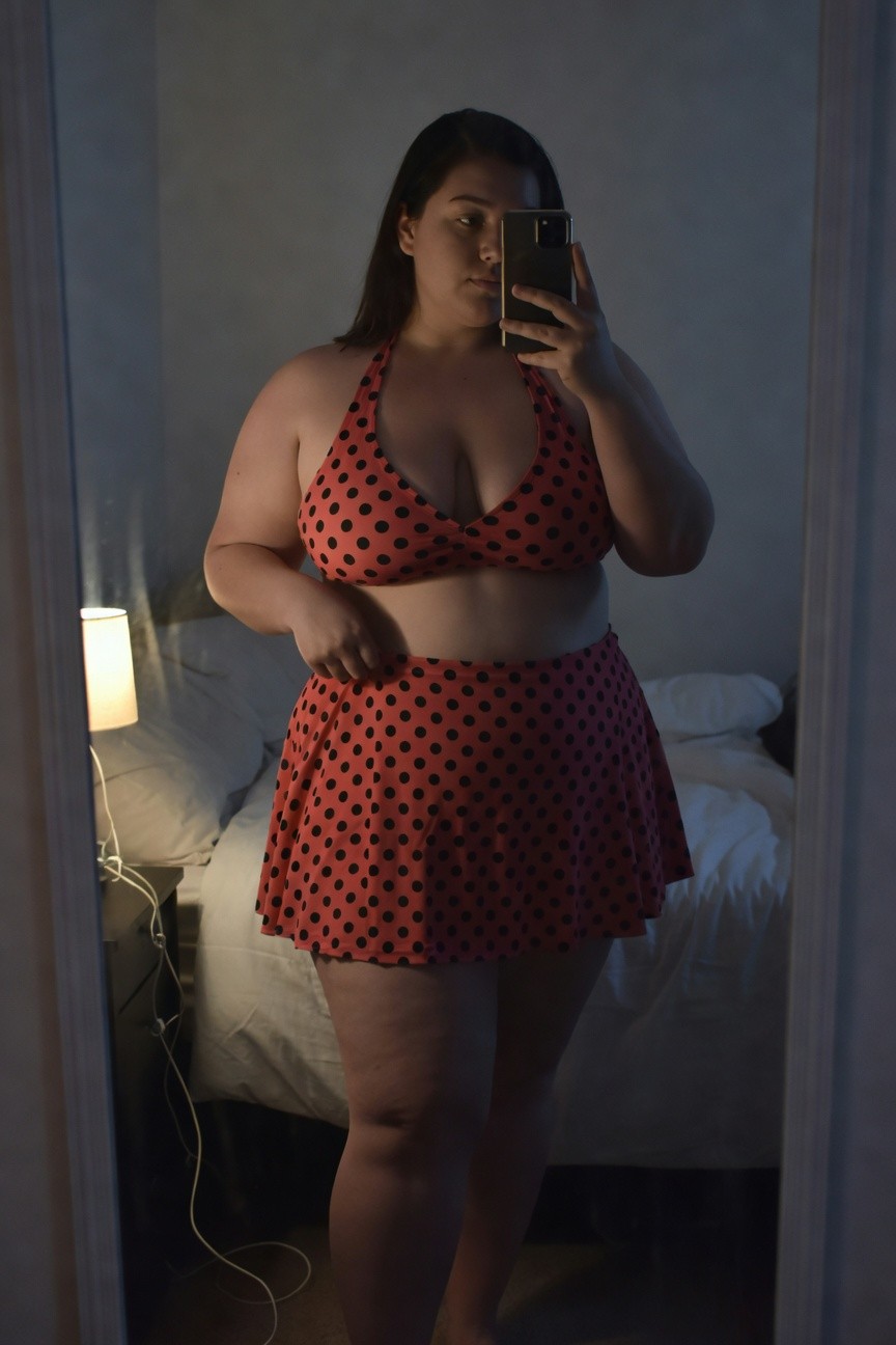 Plus-size woman with long dark hair taking a mirror selfie in a red halter bikini top with black polka dots and matching high-waisted flared polka dot skirt, standing in a bedroom with bed and lamp visible
