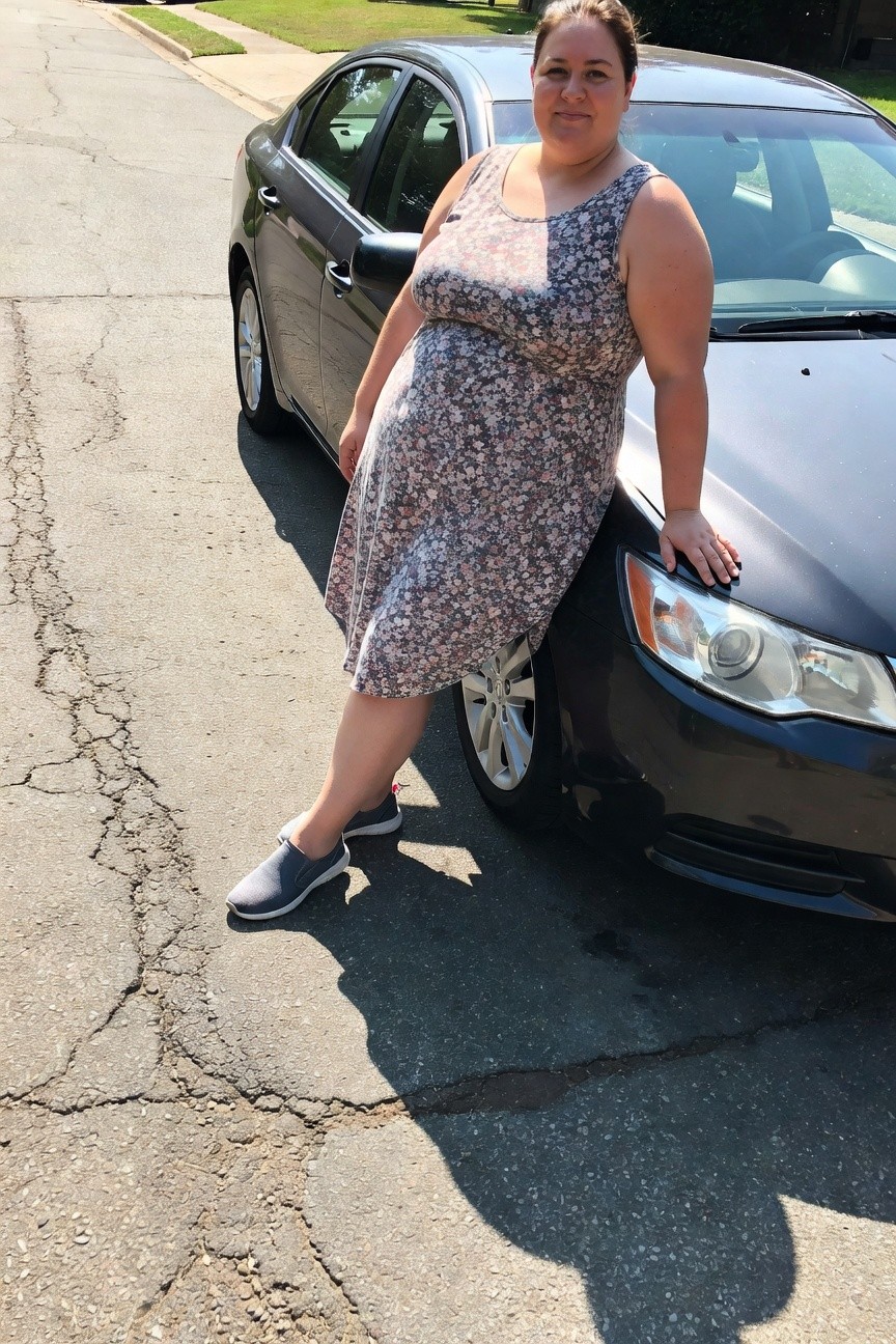 Plus-size woman in gray-based floral sleeveless midi dress and gray slip-on sneakers, leaning against a black car on a sunny street