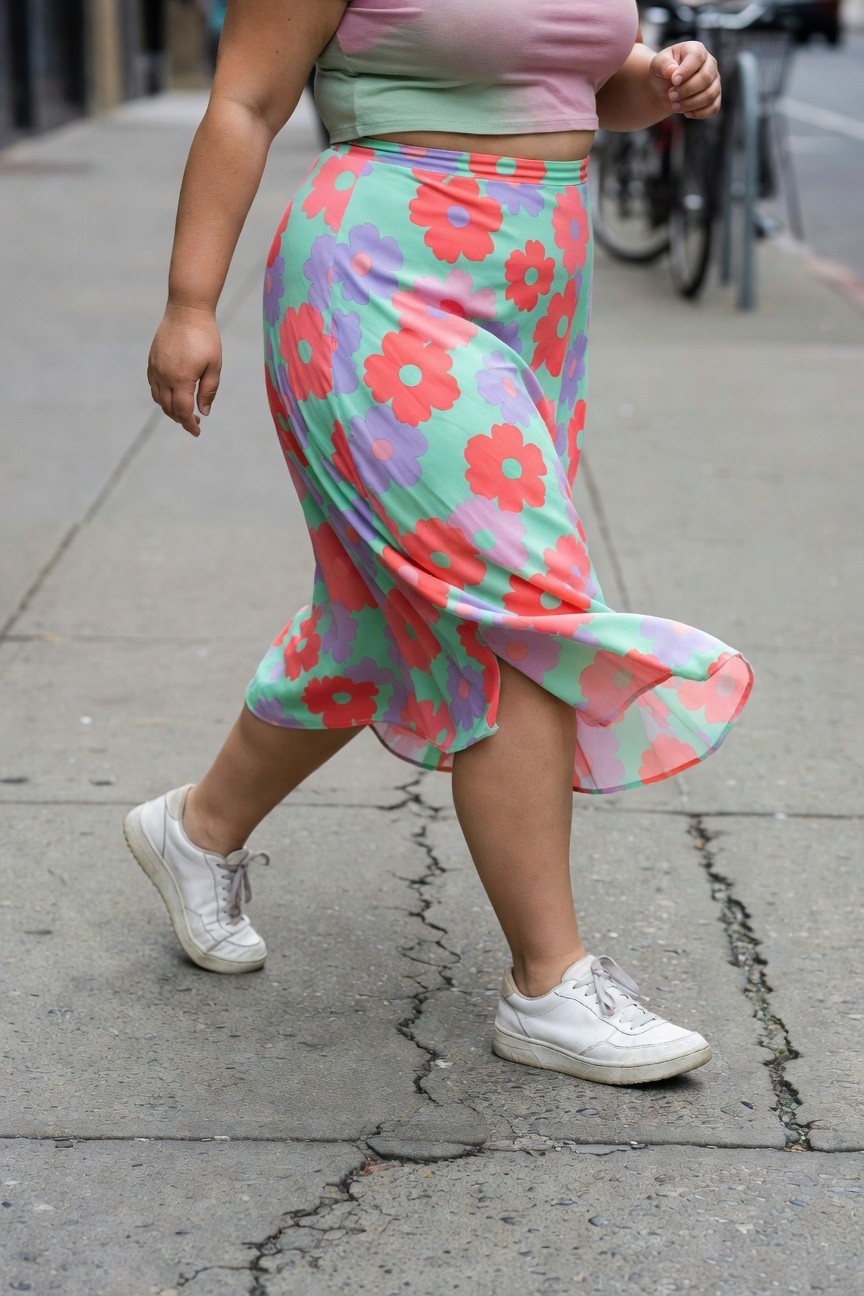 Plus-size woman in a flowy multicolored floral print skirt with pink crop top and white sneakers, captured mid-stride on a sidewalk