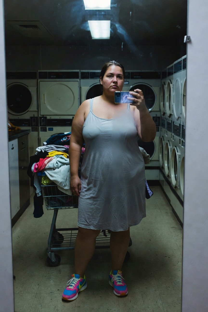 Plus-size woman taking a mirror selfie in a laundromat, wearing a loose gray tank slip dress and bright pink Nike sneakers with colorful accents, laundry cart nearby